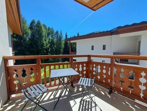 Een balkon met een tafel en stoelen, een houten hekwerk en een uitzicht op bomen en een gebouw op de achtergrond.