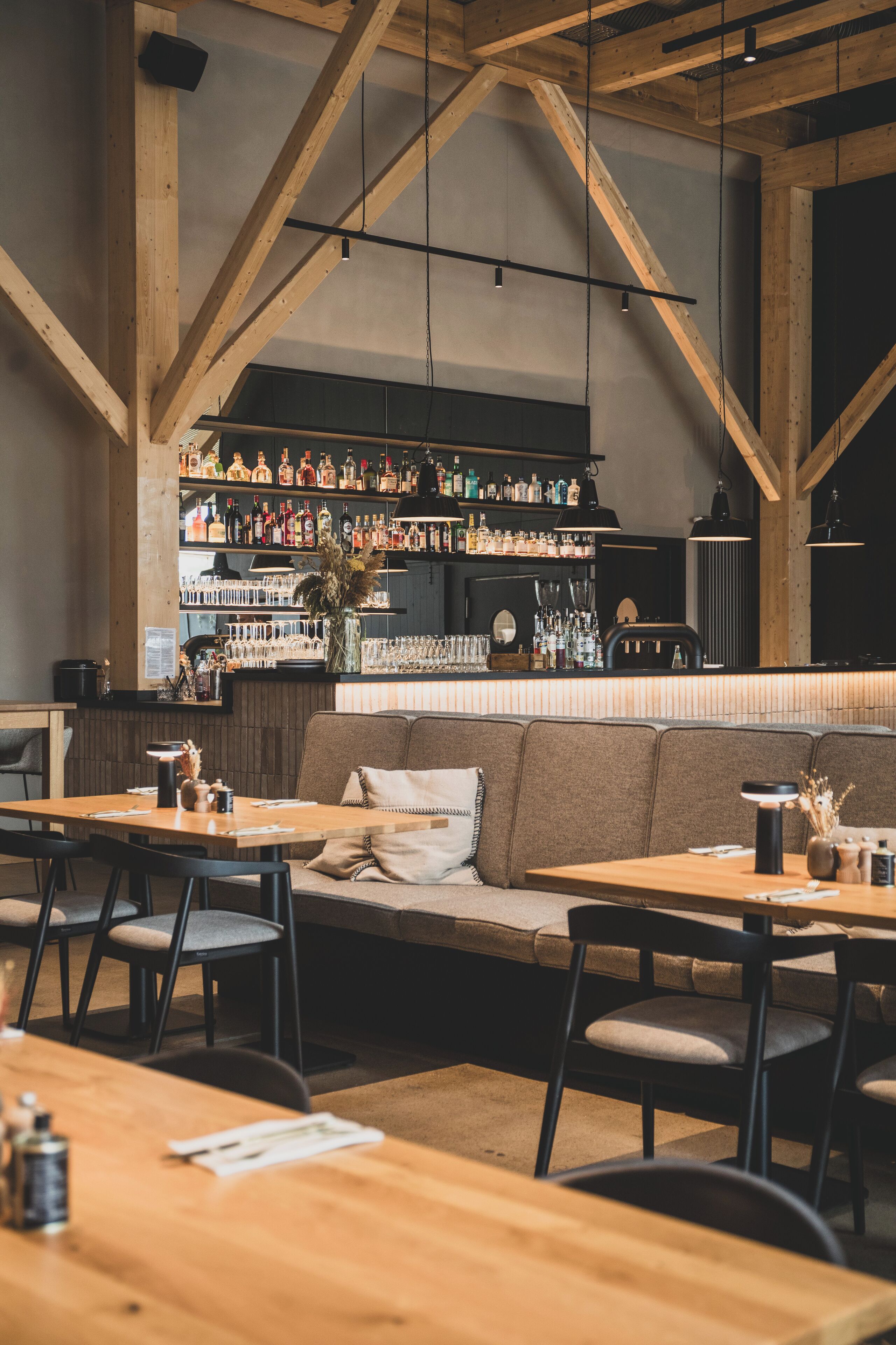 Interior de restaurante con vigas de madera, zona de bar con estantes llenos de botellas y área de comedor con mesas y sillas.