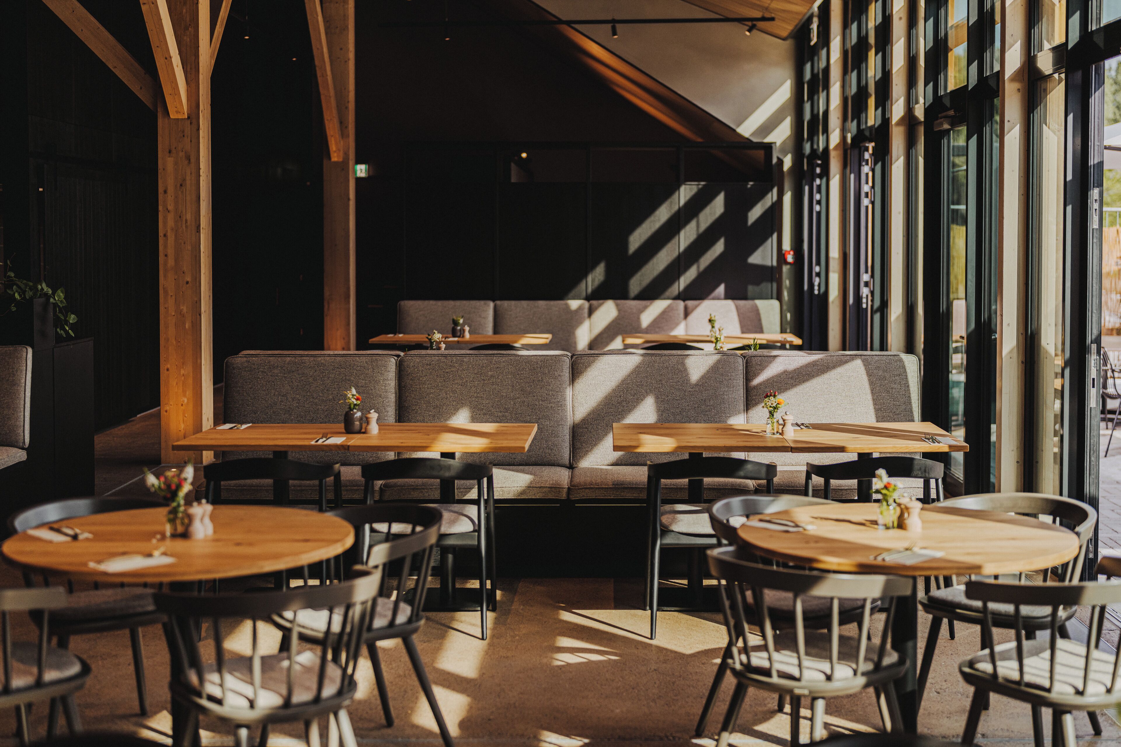 A cozy cafe with wooden tables and chairs, a cushioned bench, and large windows allowing natural light.