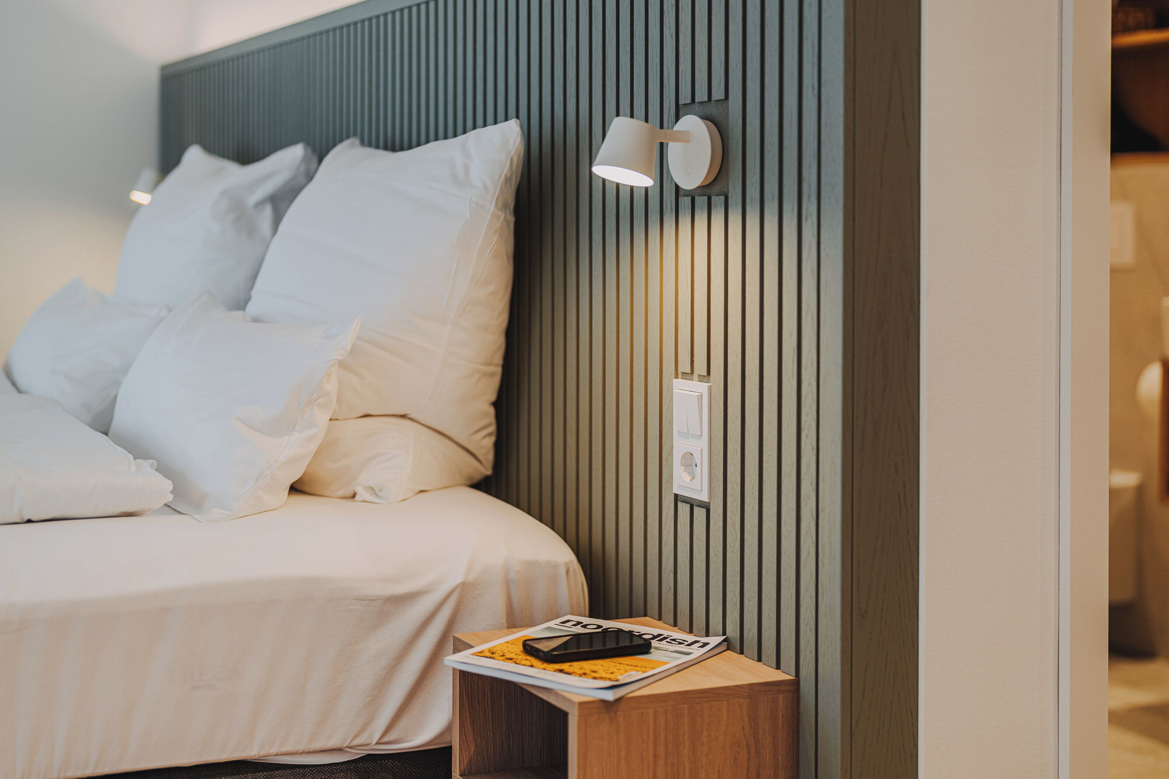 A neatly made bed with white linens and pillows, a bedside table with a magazine and a smartphone, and a wall-mounted light fixture.