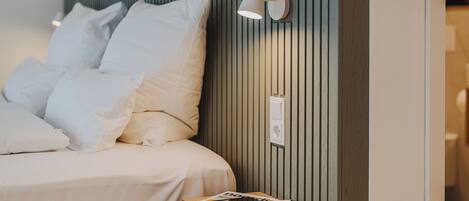 A neatly made bed with white linens and pillows, a bedside table with a magazine and a smartphone, and a wall-mounted light fixture.