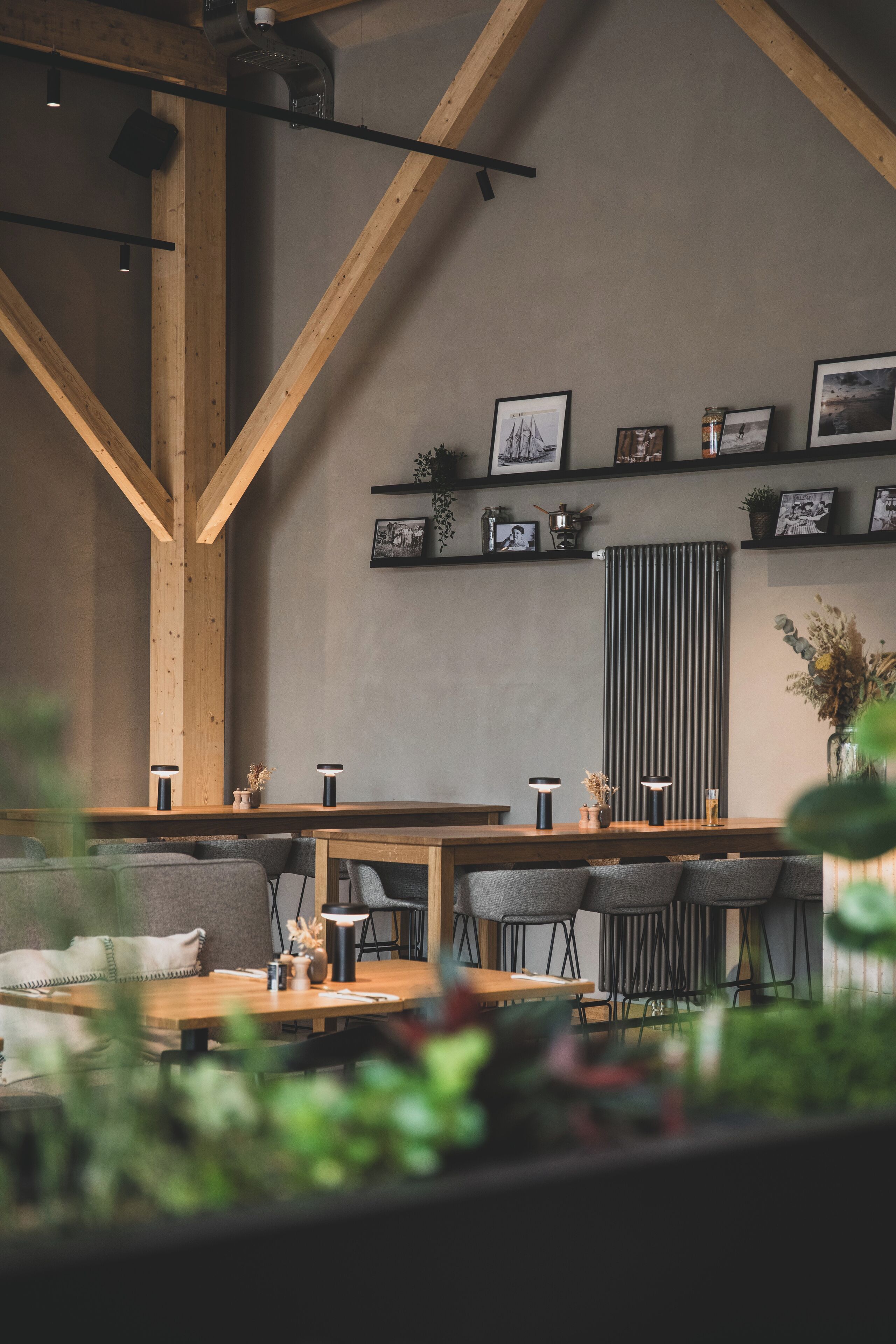 Ein gemütliches Café mit Holzbalken, einem langen Tisch und Stühlen. An der Wand hängen gerahmte Bilder und ein Heizkörper ist zu sehen.