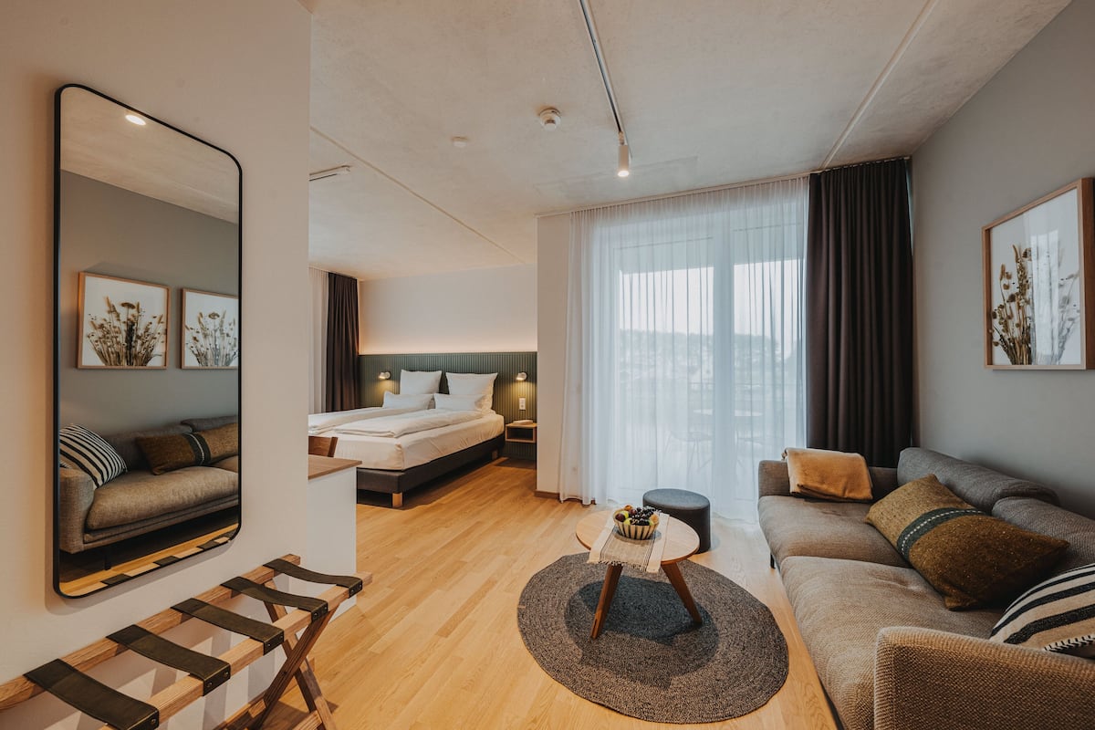 A modern hotel room with a bed, sofa, and a dining area.