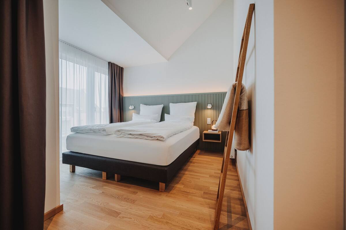 A modern hotel room with a large bed, wooden flooring, a sloped ceiling, and a view of the city through large windows.