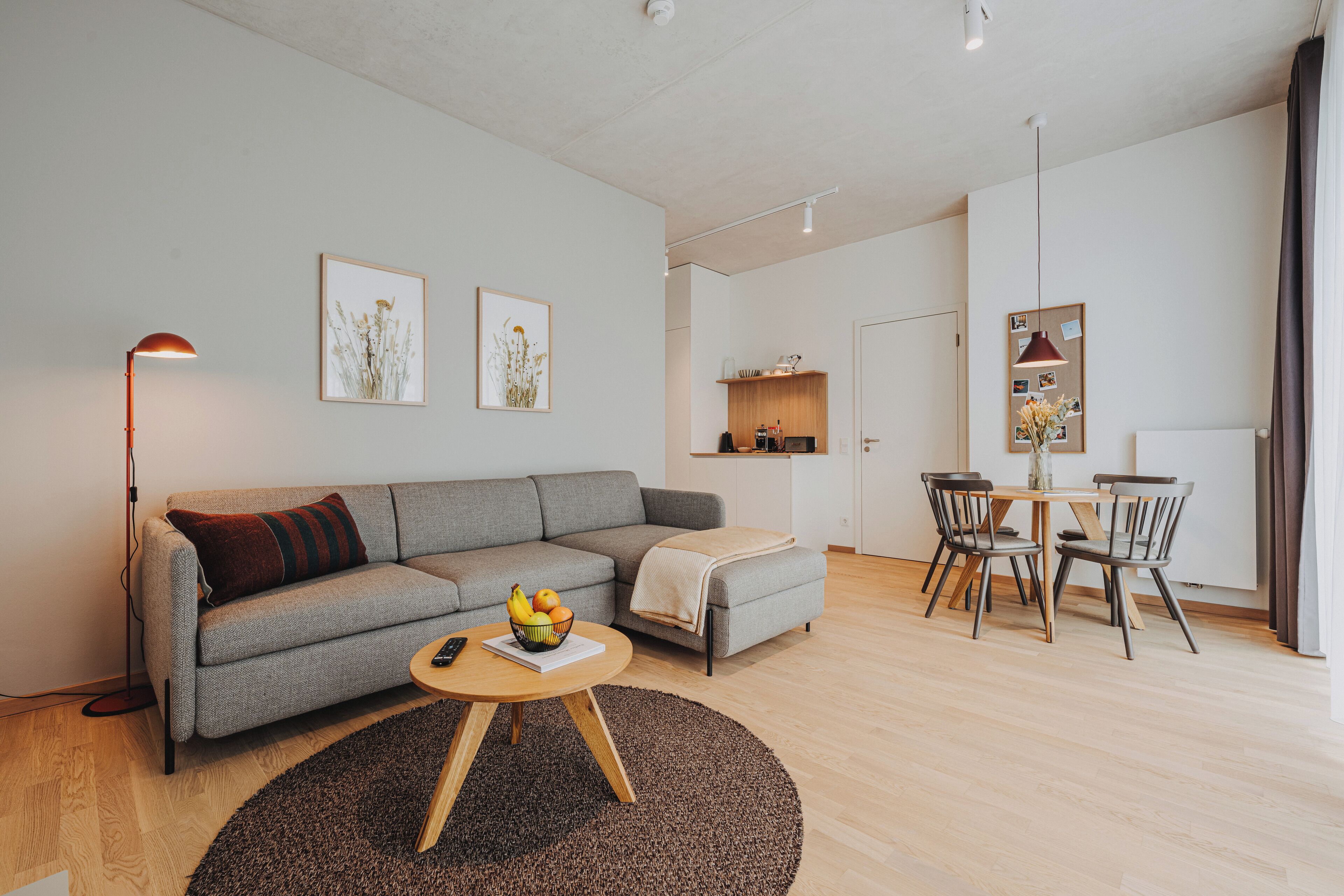A modern living room with a grey sofa, a round wooden coffee table with fruits, a floor lamp, and a dining area with a small table and chairs.