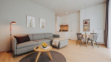 A modern living room with a grey sofa, a round wooden coffee table with fruits, a floor lamp, and a dining area with a small table and chairs.