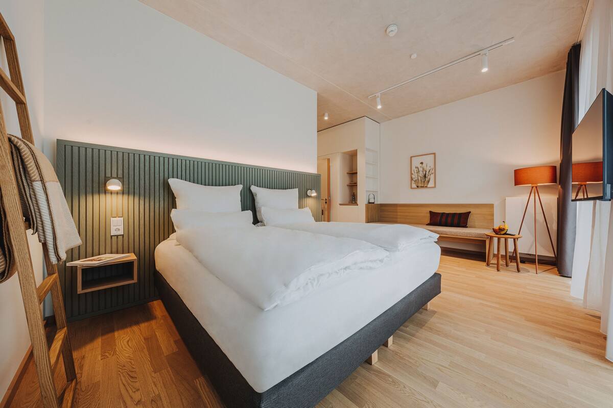 A modern hotel room with a large bed, a bedside table, a small table with a lamp, and a view of the hallway.