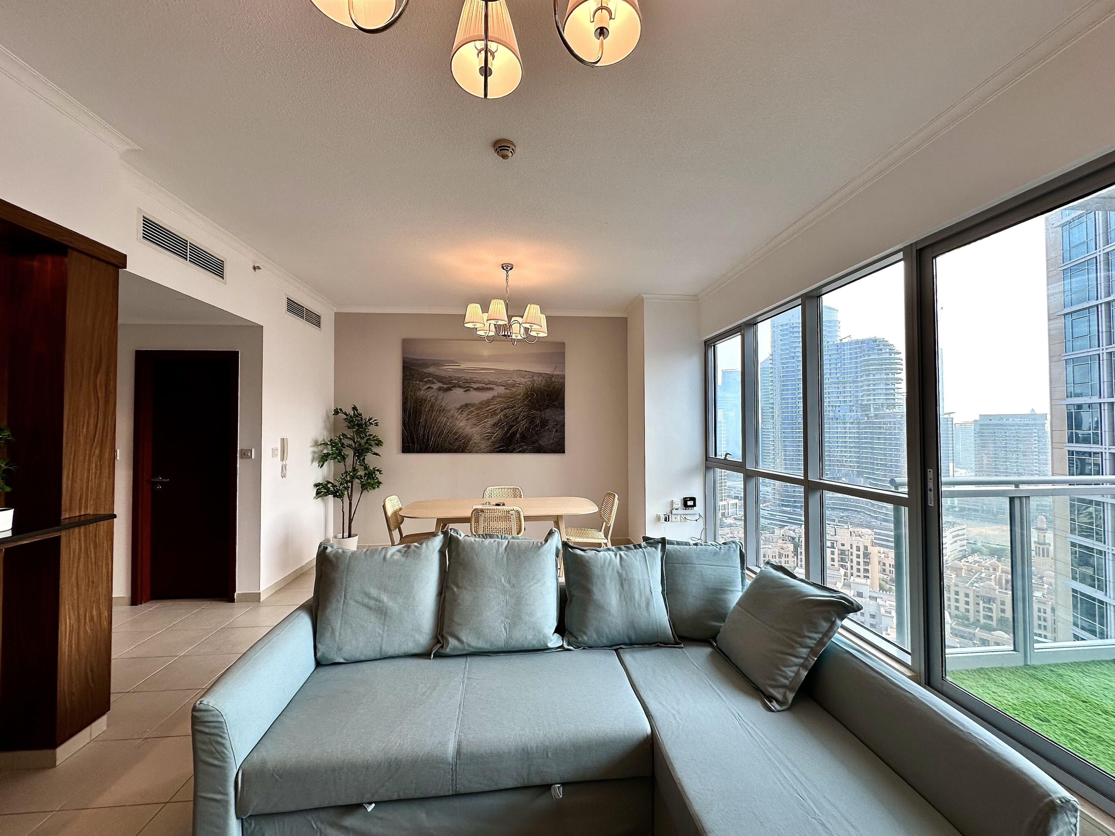 A modern living room with a sectional sofa, a dining table with chairs, a large window with a city view, and a chandelier.