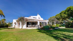 Eine weiße Villa mit Terrasse, umgeben von einem gepflegten Rasen und unterschiedlichen Pflanzen.