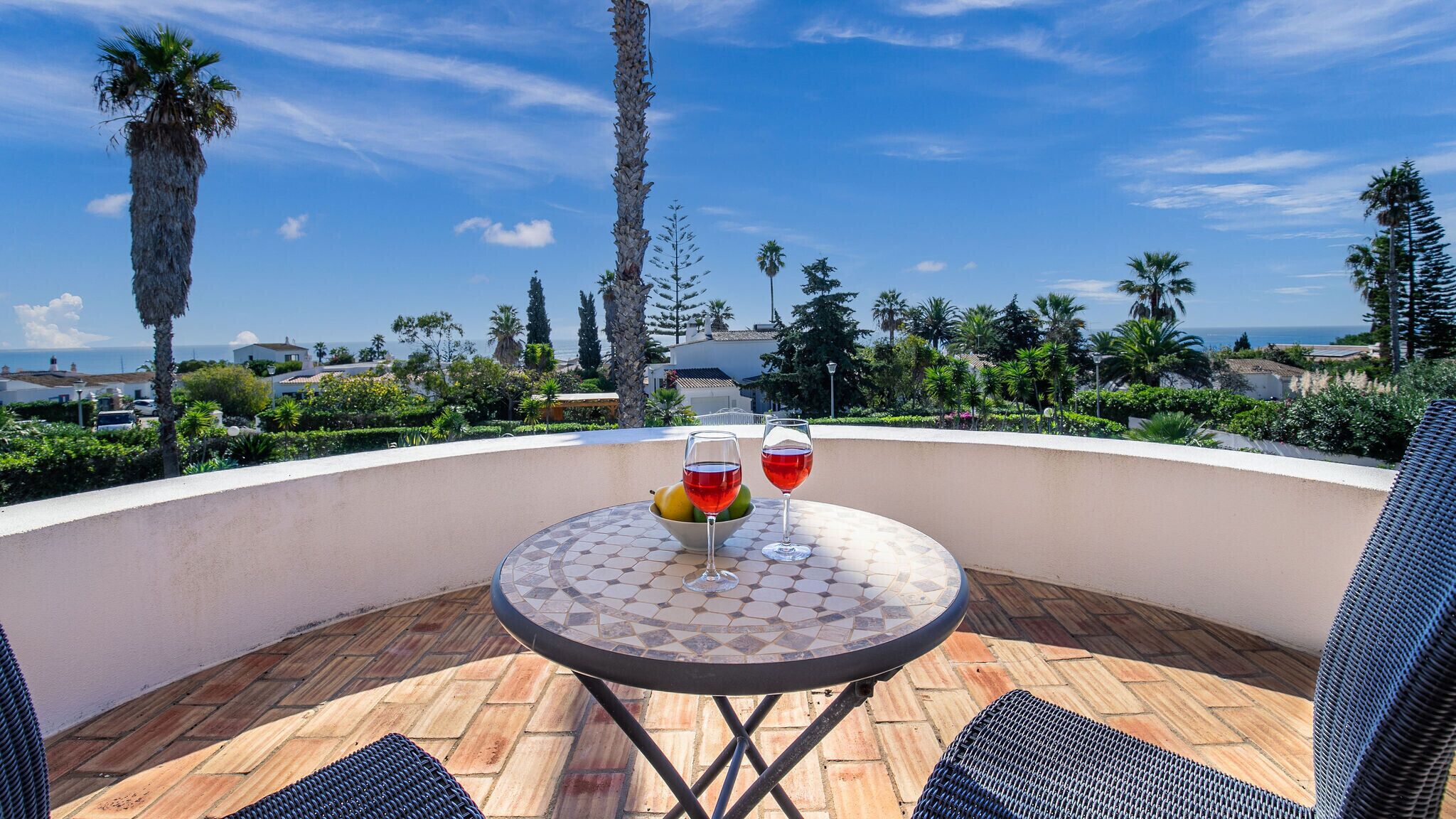 A patio with a round table, two glasses of red wine, and a bowl of fruit.