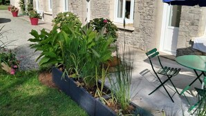 A stone building with a garden area featuring a raised planter bed with various plants and a green outdoor table with a blue umbrella. - Charming house in Normandy, pets allowed, parking (Hautteville-Bocage)