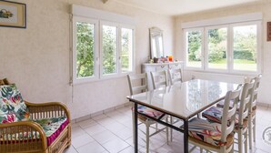 A dining area with a glass table and white chairs, large windows with a view of greenery, a wicker chair with a floral cushion, and a white cabinet with a mirror. - Charming house in Normandy, pets allowed, parking (Hautteville-Bocage)