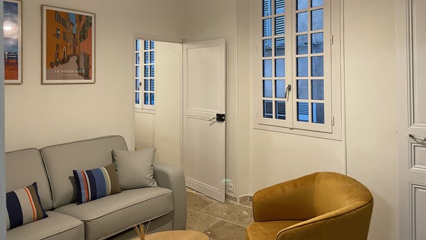 A room with a sofa, a chair, a door, and a window with shutters. - Vieux-nice 3 Pièces Confortable et Calme (Nice)