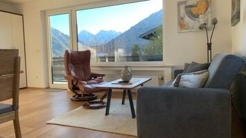 Ein modernes Wohnzimmer mit großem Fenster, einem Ledersessel, einem grauen Sofa und einem kleinen Tisch mit einer dekorativen Vase.