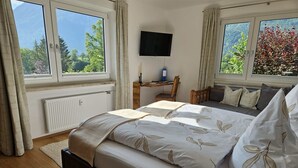 A bedroom with a bed, a desk, a chair, a TV, and large windows with curtains.