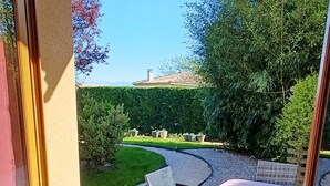 Quarto com uma grande janela com vista para um jardim ajardinado, uma cadeira e uma pequena mesa.