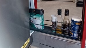 Uma cozinha com uma geladeira de aço inoxidável, uma prateleira com potes rotulados 'The Sauce' e uma caixa de papéis de filtro.