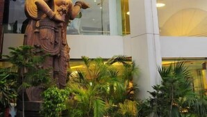 Une statue d’une divinité se trouve devant un bâtiment moderne doté de grandes fenêtres et agrémenté de plantes en pot.