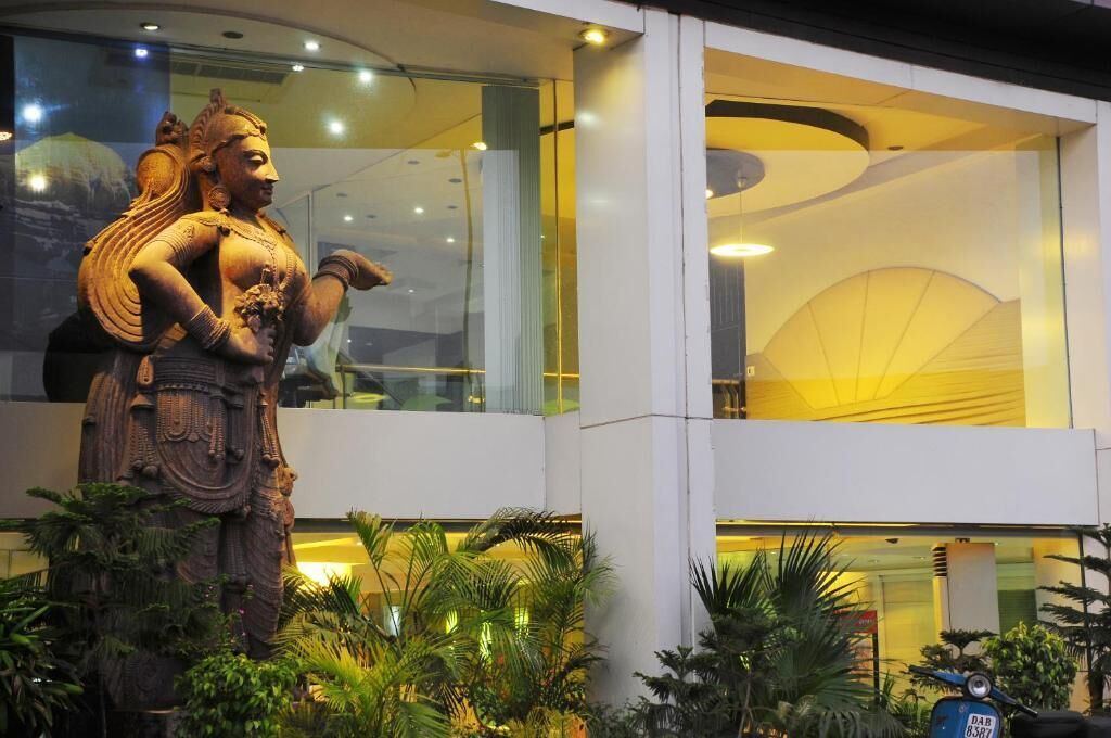 A statue of a deity in front of a modern building with glass windows and indoor plants.