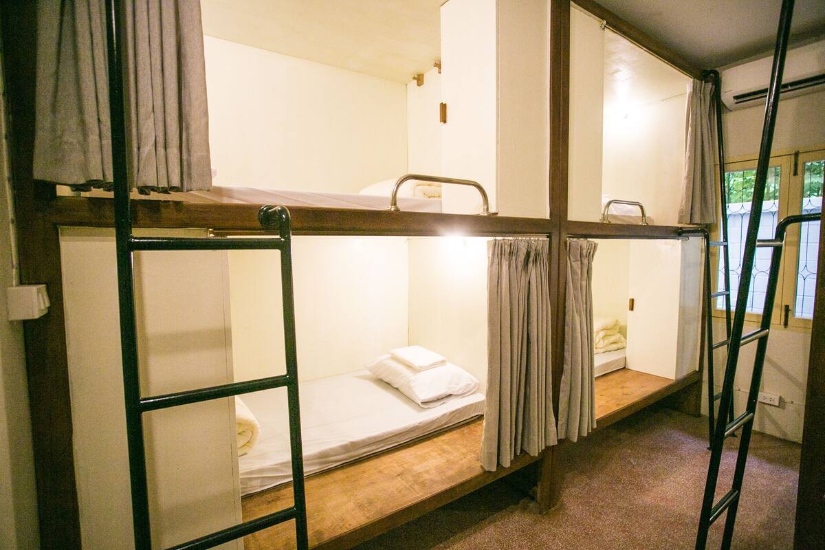A dormitory room with bunk beds, a ladder, and a window with curtains.