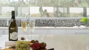A bottle of wine, two glasses of wine, a cork, grapes, and strawberries on a table with a view of trees and a tiled backsplash.