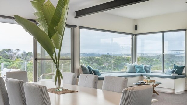 A modern dining area with a large table and chairs, a large plant, and a seating area with cushions and a view of the landscape.