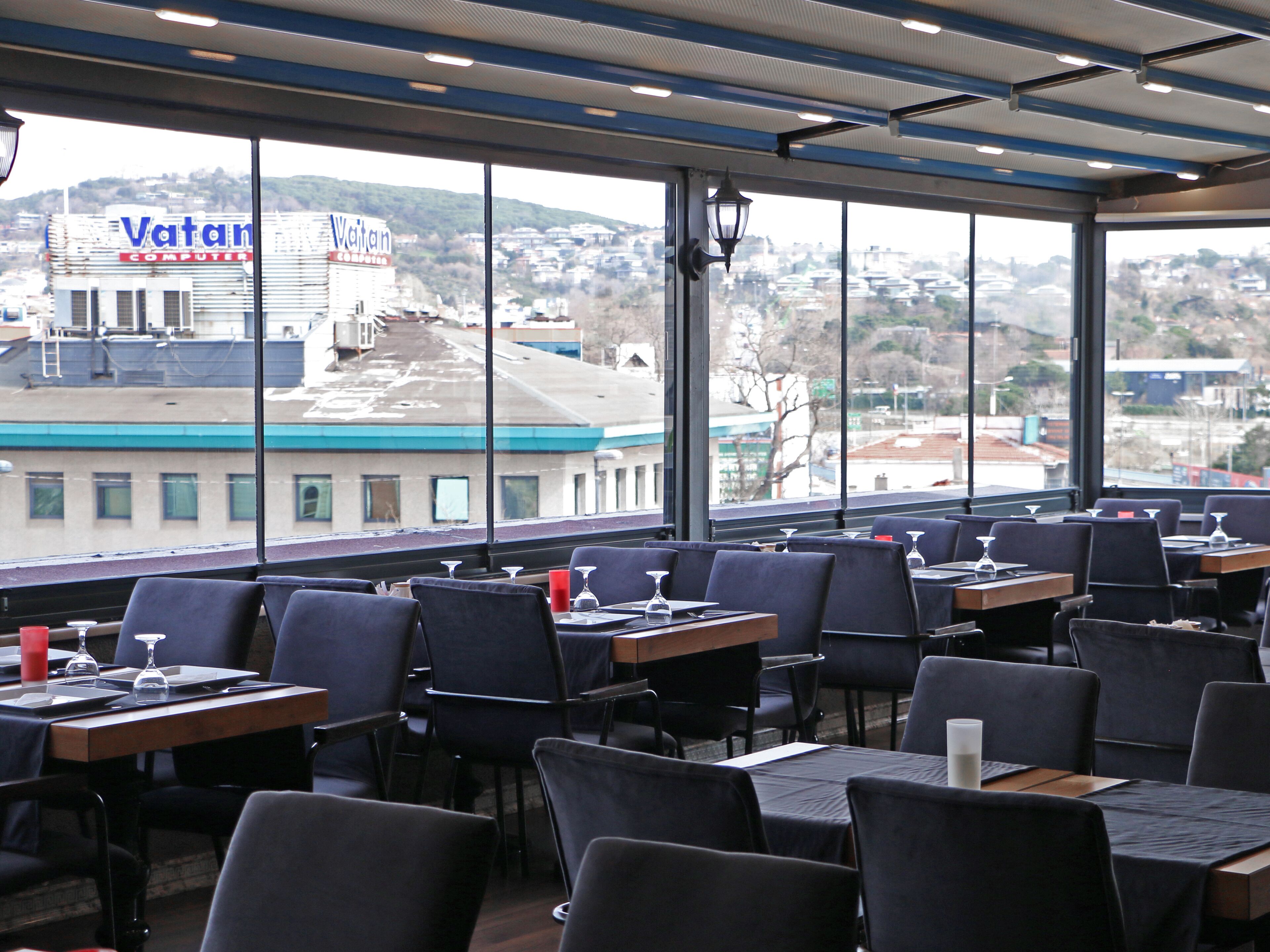 A restaurant with tables and chairs, large windows, and a view of a cityscape including a Vatan Computers building.