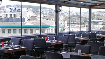 A restaurant with tables and chairs, large windows, and a view of a cityscape including a Vatan Computers building.