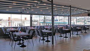 Un restaurante con un interior moderno, que cuenta con un techo alto con iluminación empotrada, amplias ventanas con vistas a la ciudad y diversas opciones de asientos, incluyendo mesas y sillas.