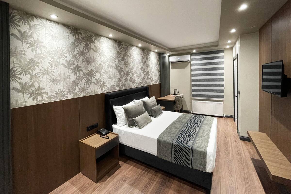 A modern bedroom with a large bed, a bedside table, a nightstand, a flat-screen TV mounted on the wall, and a window with blinds.
