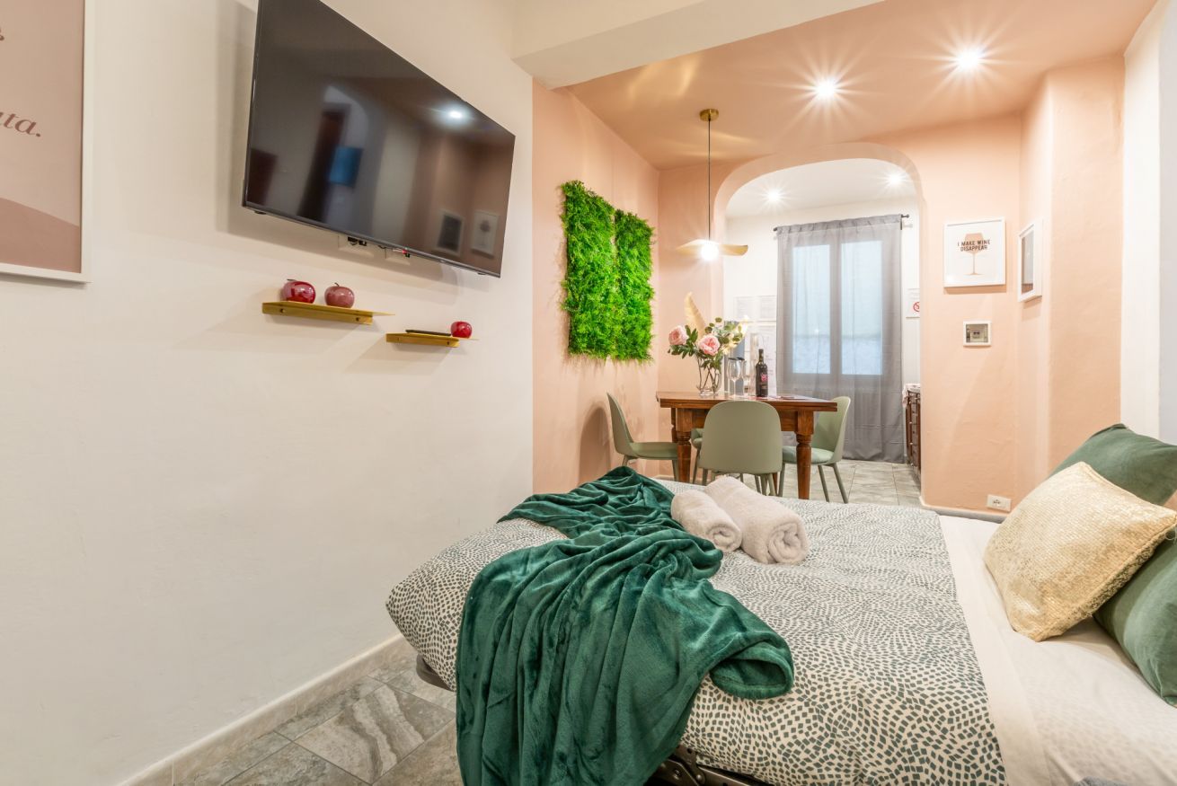 A modern bedroom with a bed, a TV, a green wall decoration, and a dining area in the background.