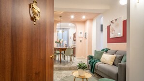 Un salon moderne avec un canapé gris, une petite table ronde agrémentée d’une plante et un pouf vert. On aperçoit à l’arrière-plan un coin repas avec une table et des chaises.