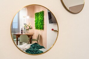 Une chambre à coucher avec un lit, une couverture verte, une chaise verte, une table en bois ornée d’un vase de fleurs et un miroir fixé au mur.