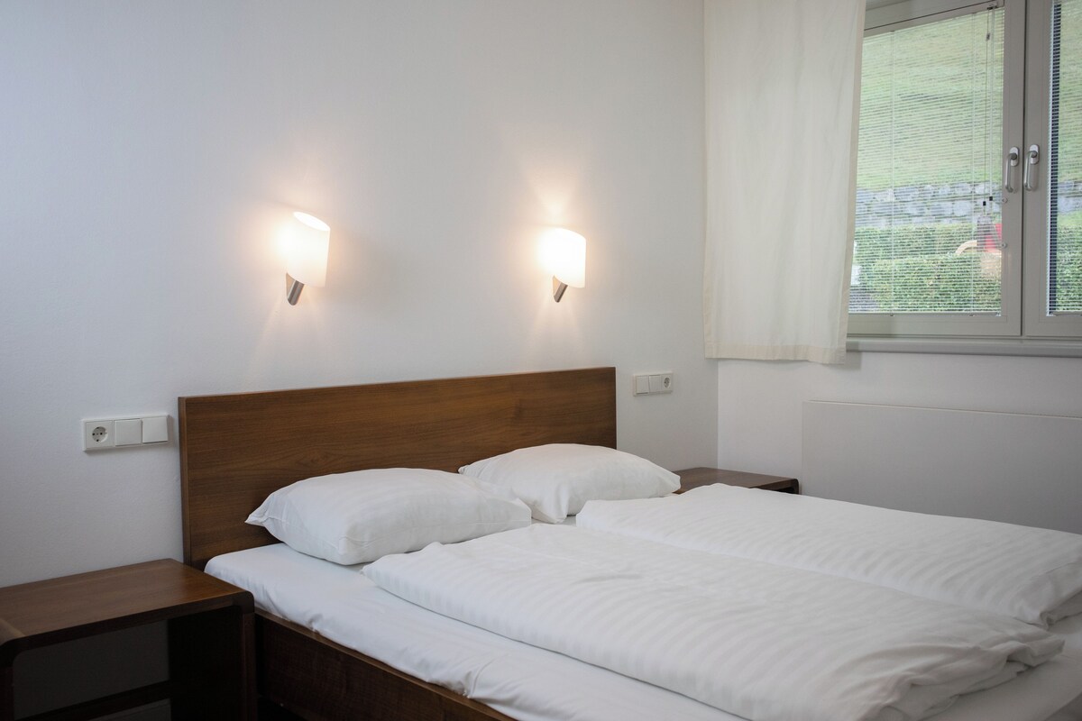 Una cama bien hecha con sábanas blancas, cabecera de madera y dos lámparas fijadas a la pared.