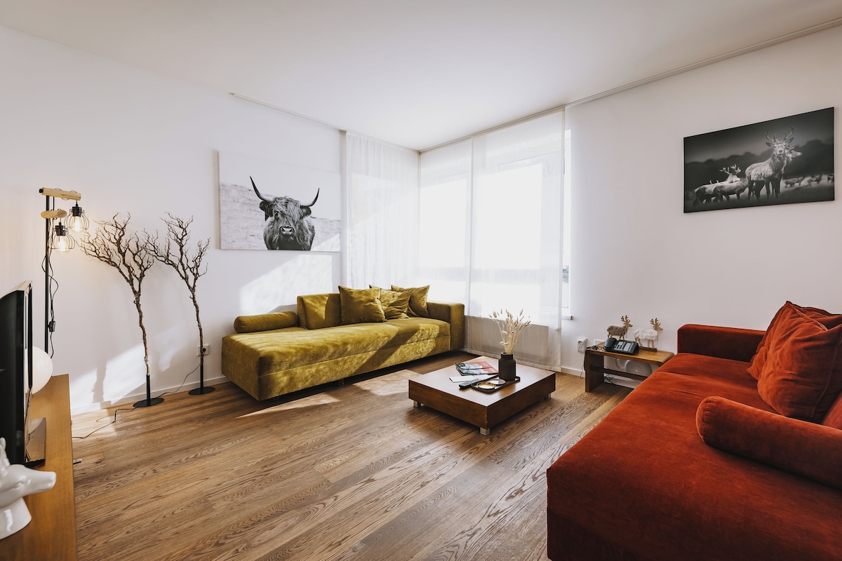Una sala de estar moderna con piso de madera, un sofá amarillo, un sofá rojo, una mesa de centro y dos lámparas de pared.