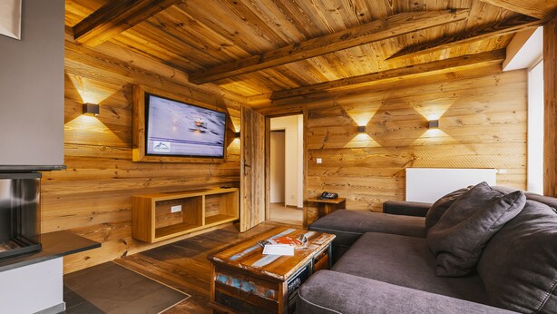 A cozy living room with wooden walls, a flat-screen TV mounted on the wall, a gray sofa, and a wooden coffee table. - Der Gletscherblick by Alpin Rentals - incl Summercard  (Kaprun)
