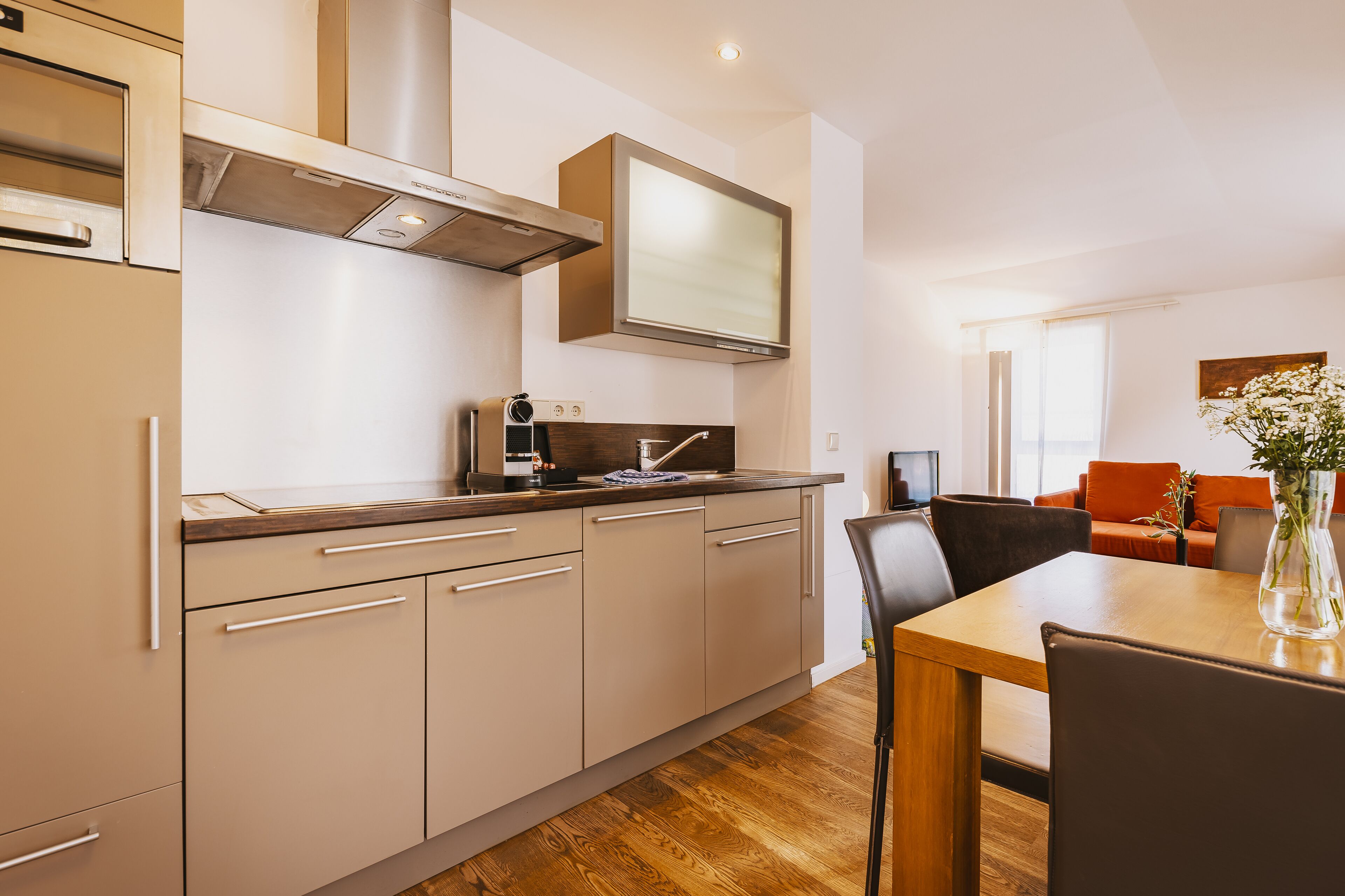 Una cocina moderna con horno empotrado, microondas y campana extractora de acero inoxidable. Cuenta con una zona de comedor con mesa y sillas de madera, y un sofá con cojines naranja.