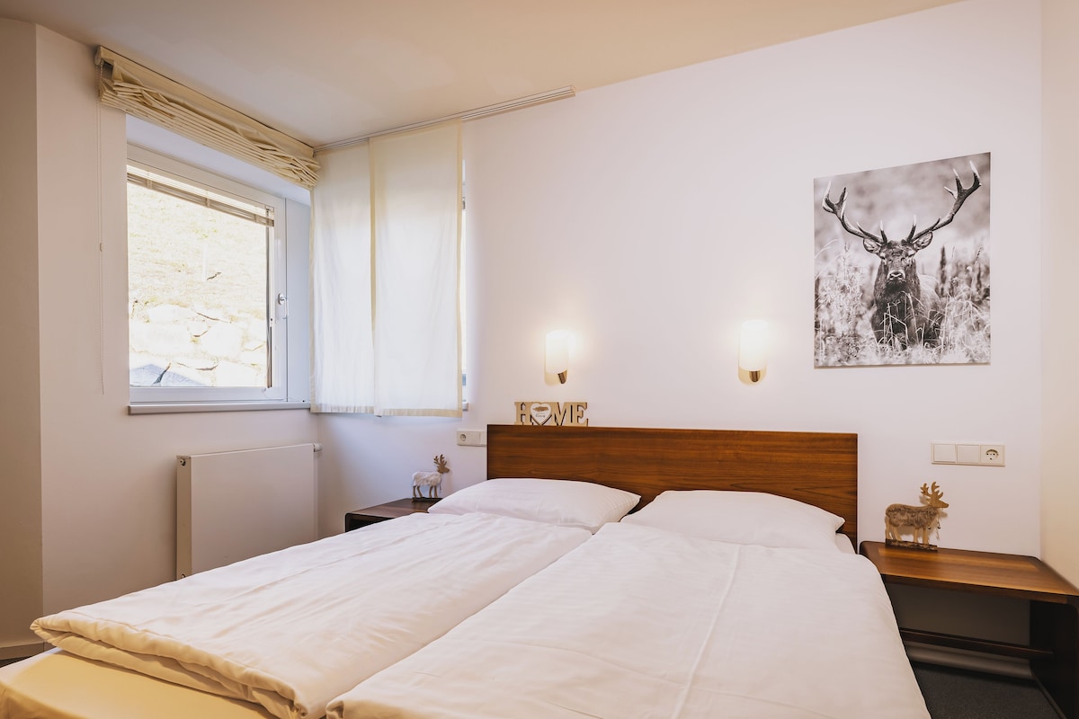 Una cama doble con sábanas blancas, cabecera de madera, mesitas de noche con lámparas, una ventana con persianas y un cuadro colgado en la pared con un ciervo.