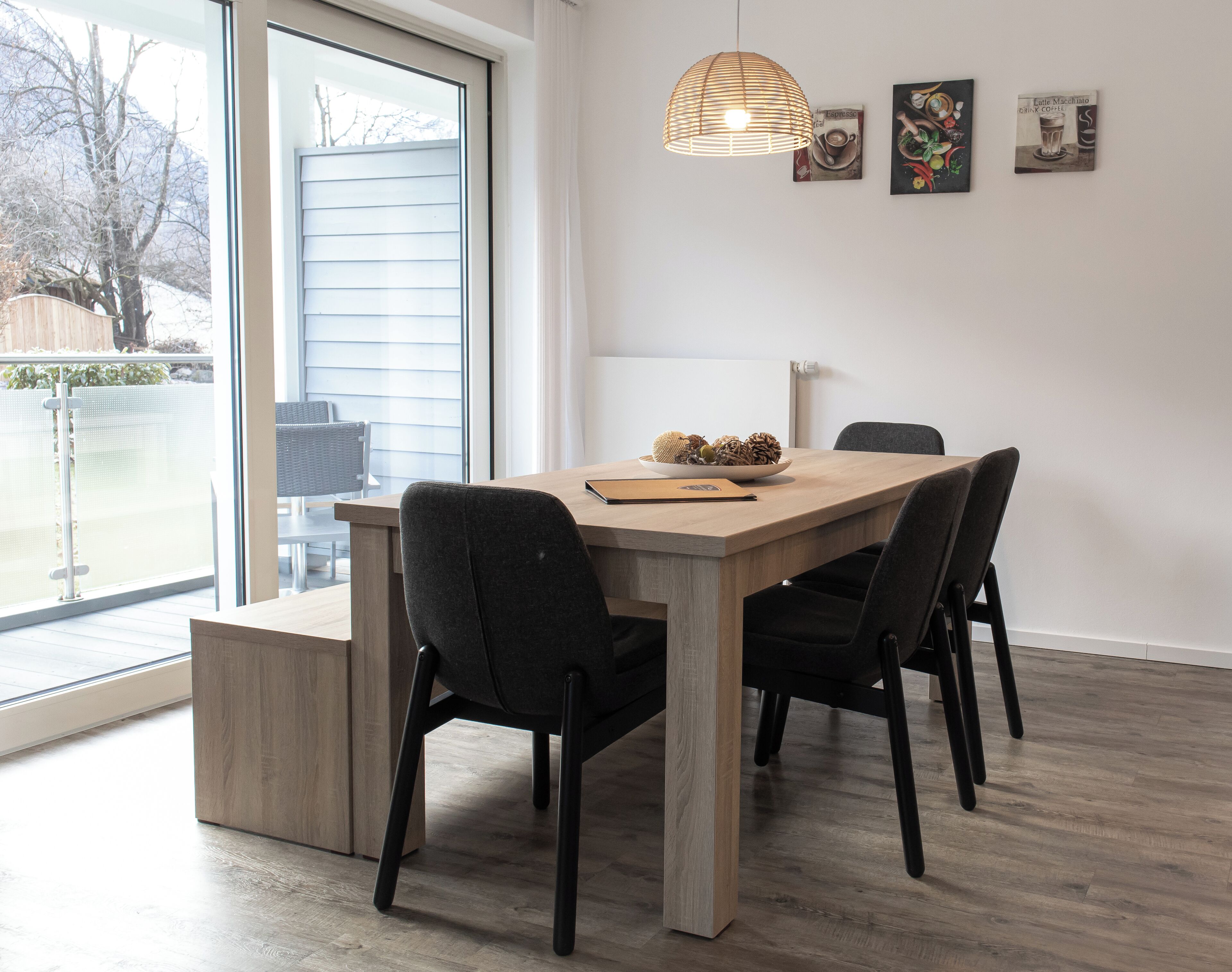 A dining area with a wooden table, four chairs, a hanging light fixture, and two framed pictures on the wall.