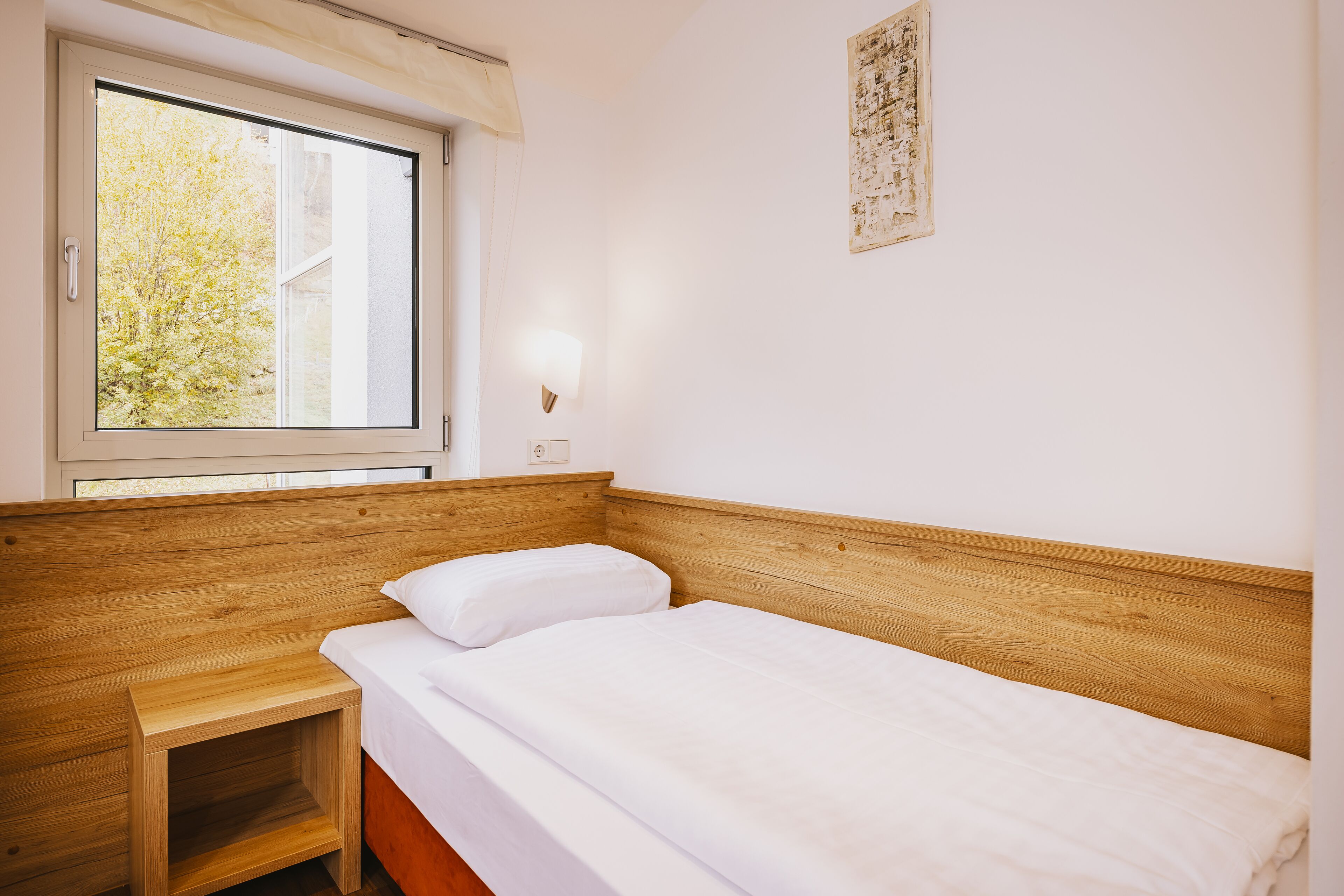 A single bed with a wooden headboard, a bedside table, a window with blinds, and a wall-mounted light fixture.