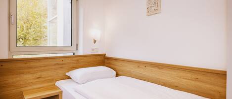 A single bed with a wooden headboard, a bedside table, a window with blinds, and a wall-mounted light fixture.