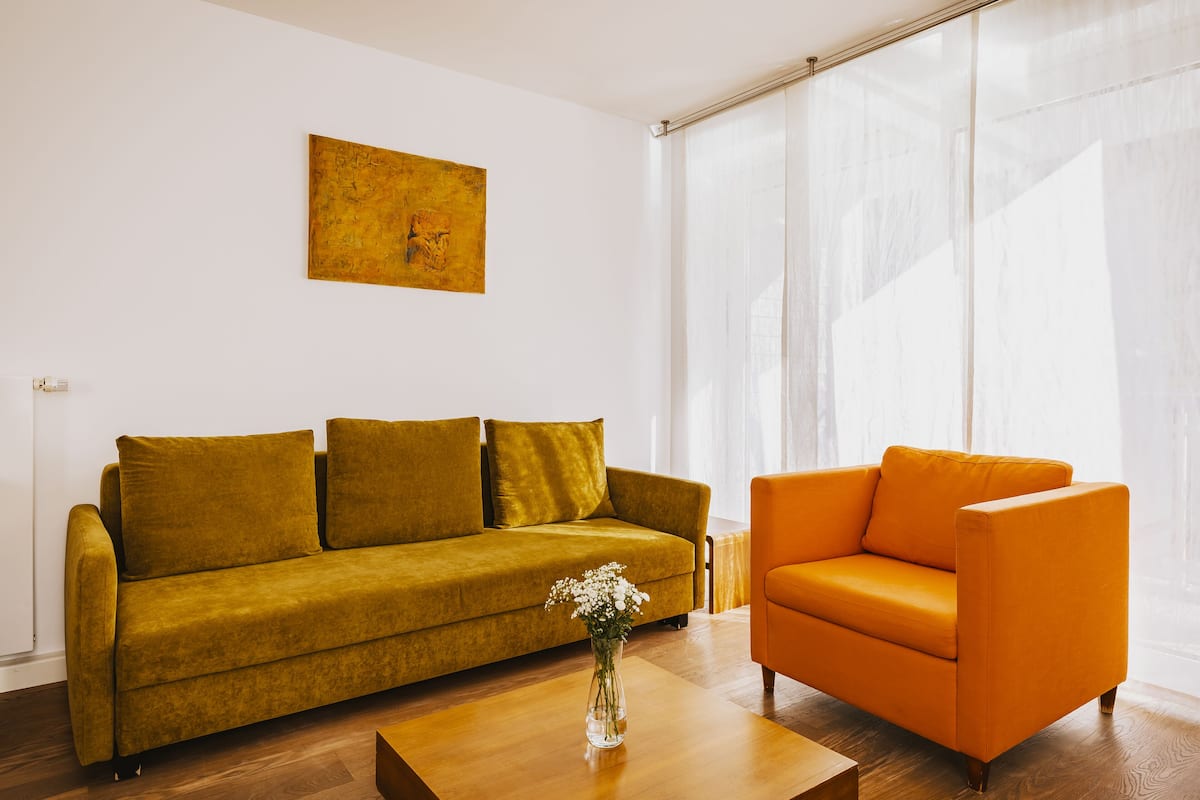 A living room with a mustard sofa, an orange armchair, and a vase with flowers on a wooden coffee table.