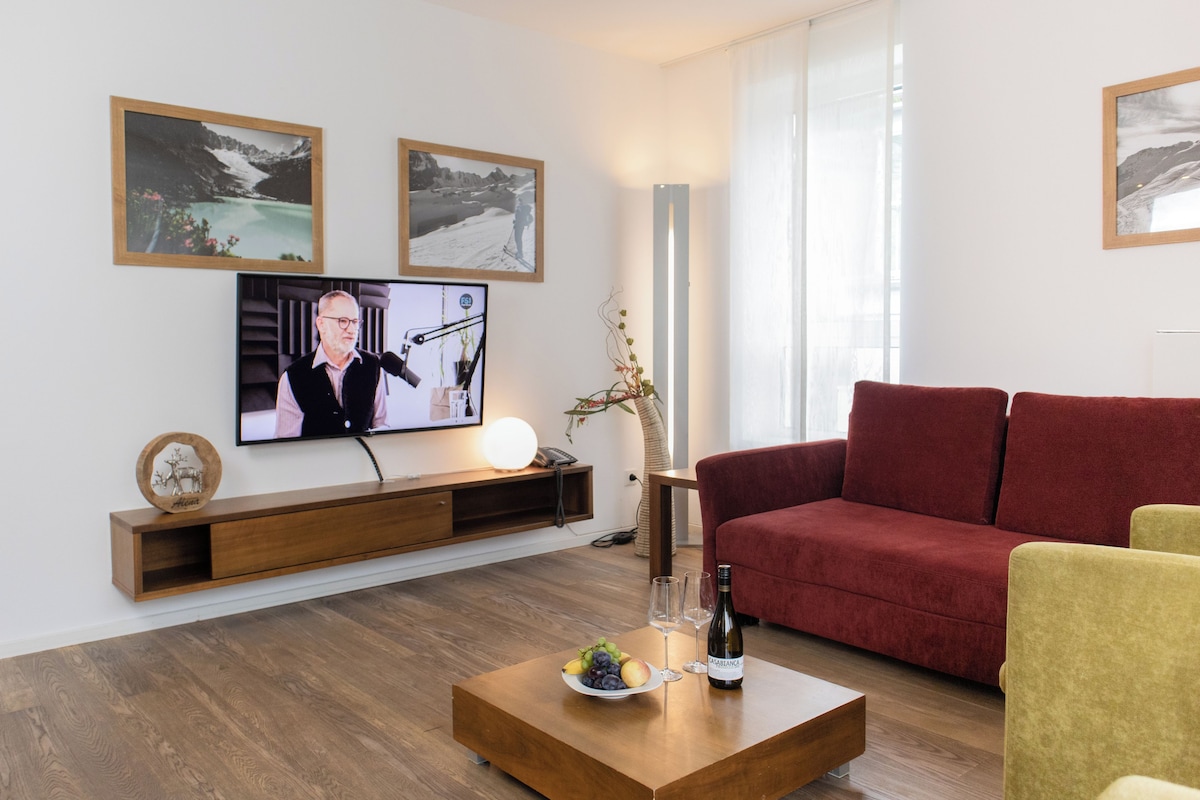 Un salón moderno con un televisor de pantalla plana, una mesa de centro de madera con fruta y una botella, un sofá rojo y un sillón amarillo.