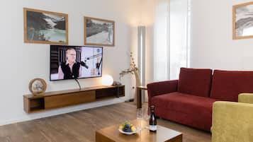 A modern living room with a flat-screen TV, a wooden coffee table with fruit and a bottle, a red sofa, and a yellow armchair.