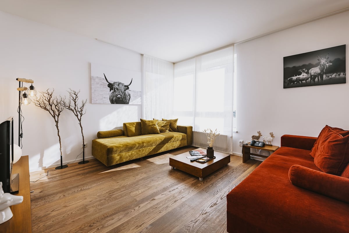 A modern living room with a wooden floor, a yellow sofa, a red sofa, a coffee table, and two wall-mounted lamps.