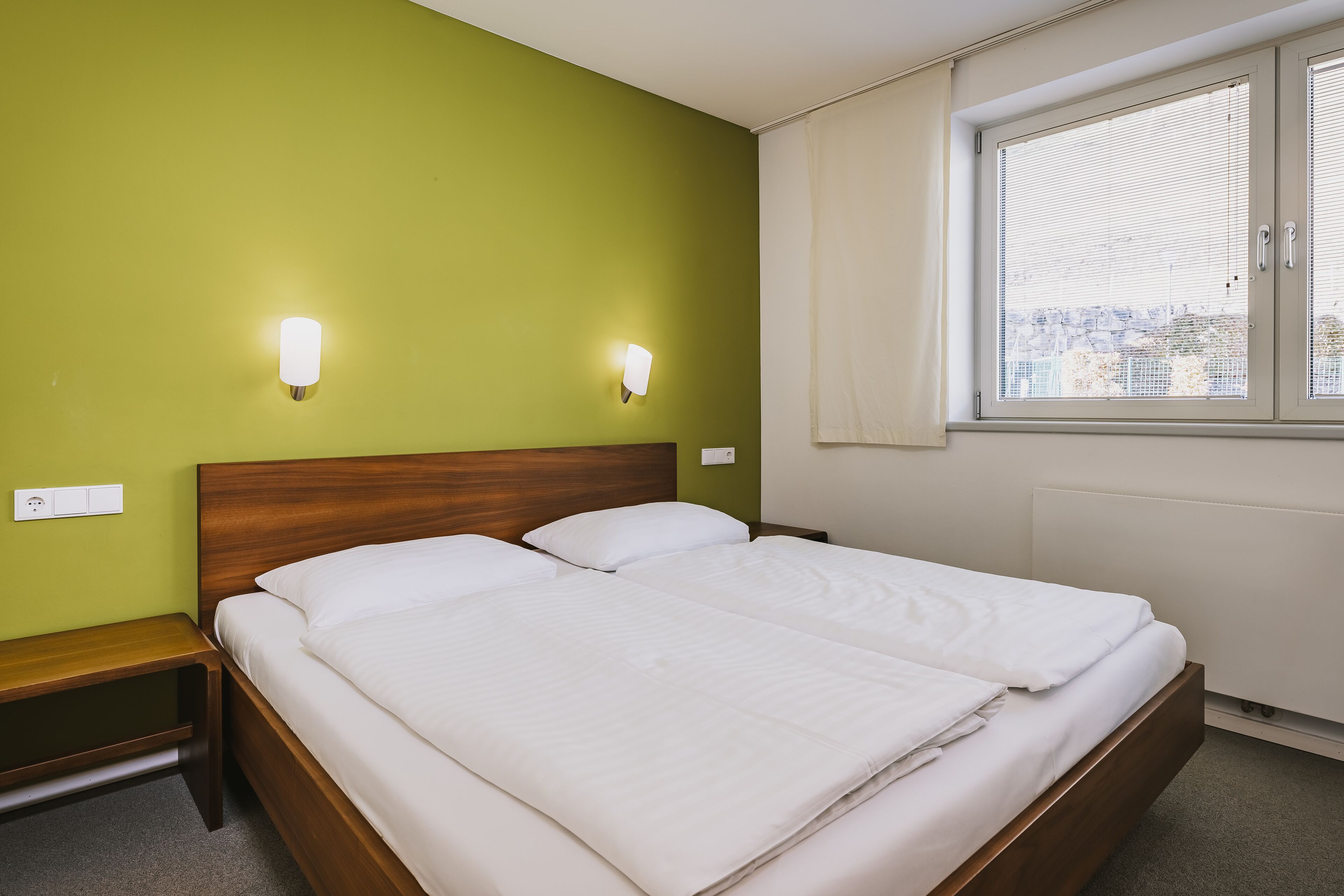 A neatly made double bed with white linens, a wooden headboard, and two wall-mounted light fixtures.