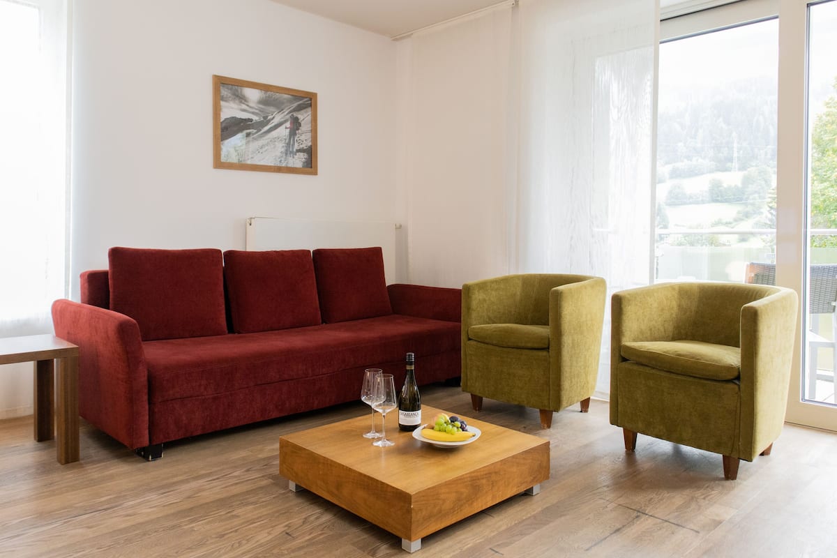 A living room with a red sofa, two armchairs, a wooden coffee table with a plate of fruit and a bottle of wine, and a painting on the wall.
