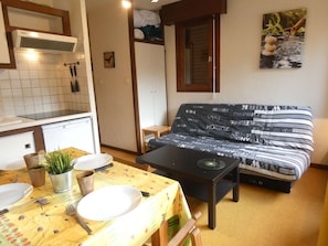 Ein kleiner Wohnraum mit Küchenzeile, einem Sofa mit gemustertem Bezug, einem Esstisch mit gelber Tischdecke und einer kleinen Topfpflanze.