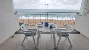 Un balcón con vista a la playa y al océano. Hay una mesa con sillas y una mesita con una taza azul y un plato.
