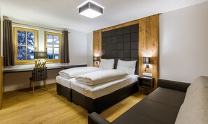 A modern bedroom with a large bed, a wooden accent wall, a window with curtains, a bedside table with a lamp, and a sofa. - Bolodges Apartments by Alpin Rentals (Saalbach-Hinterglemm)